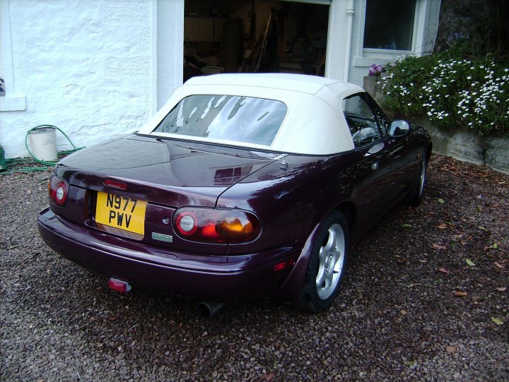 My Vr ltd comb A arrived today  - Page 1 - Mazda MX5/Eunos/Miata - PistonHeads - The image shows a convertible automobile parked on a gravel driveway in what appears to be a residential area. The car is dark-colored with a white convertible top. It has a yellow license plate with the letters "PVW" and "N137" indicated on the back. The vehicle has a modern design with distinctive tail lights and a compact shape that is typical of convertibles. The surroundings include a white building in the background with a door opening to the driveway where the car is parked.