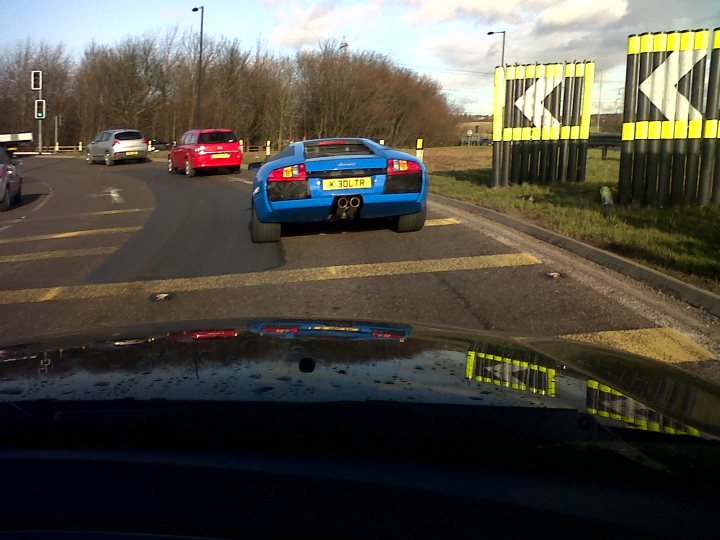 Spotted! Real Rubbish Chav Number Plates - Page 65 - General Gassing - PistonHeads - The image captures a moment on a highway, where a blue Lamborghini sports car is sitting at a traffic light on the right lane. The car's striking blue color stands out against the muted tones of the highway and the overcast sky. Behind the Lamborghini, there is a red SUV and a gray car also waiting at the red traffic light. The road is lined with grassy areas, while traffic lights are visible intermittently along the length of the highway. The overall scene suggests an overcast day with no immediate threat, allowing the driver to maintain focus on the road ahead.