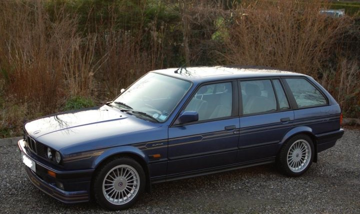 Show Me Your BMW!!!!!!!!! - Page 136 - BMW General - PistonHeads - The image features a sleek, dark-colored classic BMW car, parked at an angle on a gravel driveway. The vehicle's design suggests it is a convertible, with a soft-top roof that is currently folded down. The car's tires are black with a distinctive white sidewall design, and the vehicle exhibits a polished finish that indicates it is well-maintained. In the background, dry vegetation can be seen, hinting at a possible rural or semi-rural setting. The image itself has a color saturation that gives it a somewhat stylized, almost painterly appearance.