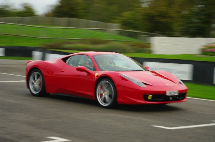 Saywell International - Page 1 - Goodwood Events - PistonHeads - The image shows a vibrant red Ferrari speeding down a road. It appears to be taken with a slight motion blur, giving the car a sense of motion. The car takes up a significant portion of the frame, indicating it is the primary focus of the image. Alongside the road is a green area, which contrasts with the road itself which is a smooth grey surface. On the right side of the image, there is a white fence, adding to the landscape. The scene suggests a fast-paced journey, likely taken during the daytime under clear weather conditions.