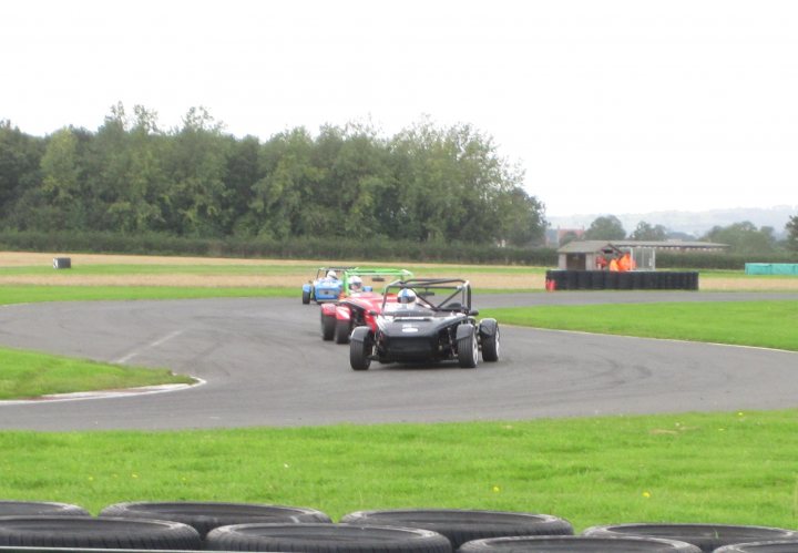 3 MEV EXOCETS RACING AT BRANDS HATCH - Page 1 - Kit Cars - PistonHeads - The image captures an exciting scene of a go-kart race taking place on a track made of tires laid down on grass. Three go-karts, each colored differently, are in mid-motion, rounding the left side of the track. The racers are wearing full racing gear, indicating a high-speed event. In the backdrop, a spacious open field can be observed, with a fence and a solitary figure in an off-road vehicle, possibly a support crew member or another participant waiting their turn. The sky overhead is overcast, casting a soft, diffuse light on the scene.