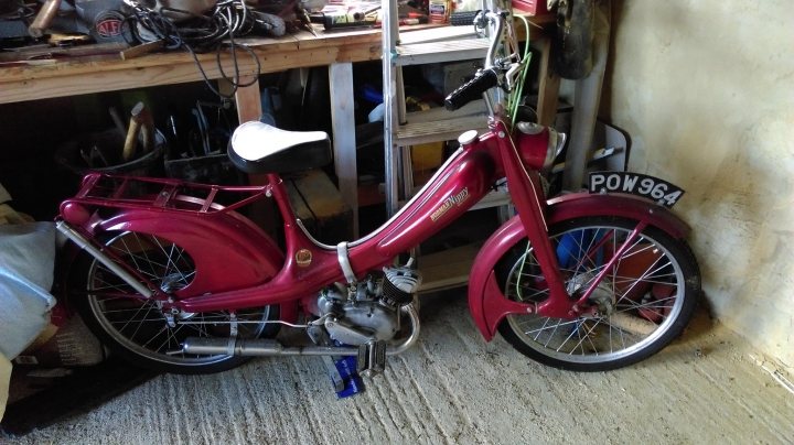 Anyone with a Holden interested in shooting a video? - Page 1 - HSV & Monaro - PistonHeads - The image features an old-fashioned, eye-catching, shiny red scooter parked on a concrete floor. The scooter's vibrant red color stands out against the more muted background. The scooter is leaning against a sturdy metal rack, suggesting possibly outdoor or industrial use. To the right of the scooter, there is a wooden shelf containing various objects, further hinting at a workshop or garage setting. Overall, the image gives an impression of a well-used vehicle in a working environment.