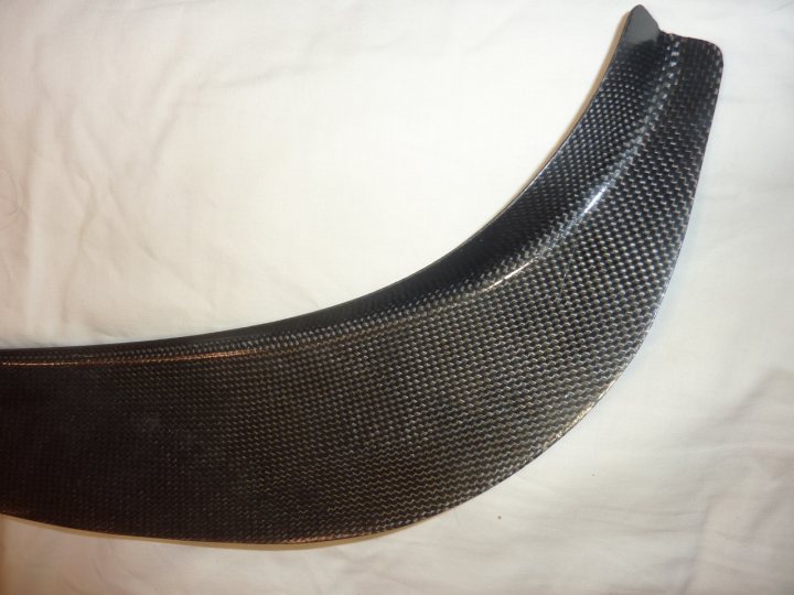 Best way to attach front dive planes? - Page 1 - Radical - PistonHeads - The image features a close-up of a curved, black object with a rough texture. It appears to be a piece of motorcycle bodywork, a carbon fiber component typically used for enhancing the aesthetics and performance characteristics of the vehicle. The carbon fiber appears to have a weave pattern, which is a characteristic of the material. The background is a textured, light-colored surface, providing a contrast to the darker tones of the object. The style of the image is straightforward and without additional context gives the impression of a random cropping to focus on the object.