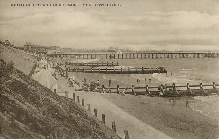 Lowestoft - Page 2 - East Anglia - PistonHeads - This image is a historical black and white photograph depicting a bustling beachfront scene with people and vehicles, likely from the late 19th to the early 20th century, as suggested by the attire and infrastructure. The foreground features a pier with buildings and people, typical of a seaside resort. In the background, vessels are visible in the water, indicating the importance of maritime activity in this coastal town. Text on the image confirms that it was taken in South Cliffs and Claremont Pier, Lowestoft. The presence of textured elements and the grainy quality are indicative of historical photography processes of the time.