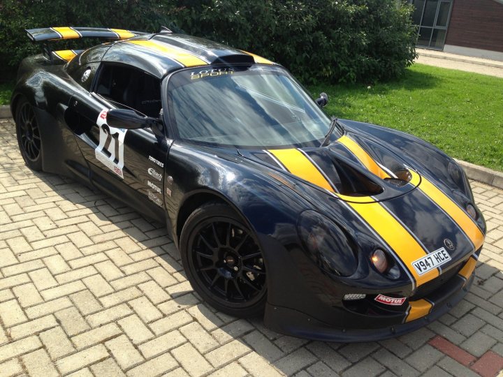 My Elise Exige 293bhp K20.  Do you know her ? - Page 1 - Elise/Exige/Europa/340R - PistonHeads - This image showcases a sleek, black sports car parked in a lot. The vehicle is adorned with striking yellow stripes on its front hood and a yellow side stripe. Notably, it displays the number 56 and the Pirelli brand of tires. The car's design suggests a fast, high-performance model. The tail end of a person is visible in the foreground, giving a sense of scale to the car's size. The car is cast against a backdrop of green grass and a brick-paved lot, which adds to the overall appeal of the vehicle.