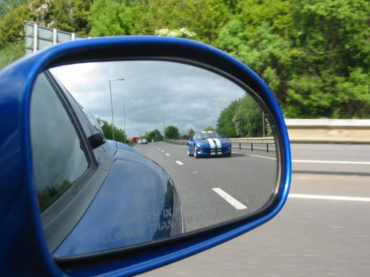 The image captures a scene inside a car, viewed through the rearview mirror. The dominant color palette of the car's interior is blue, with reflective door panels visible in the mirror. Through this mirror, the reflection captures a slice of life on a highway, where a blue sports car with white stripes is visible. The car is ahead and moving from the front end of the car in mirror. Other vehicles, including a red truck and a white van, are captured further down the road, suggesting a busy and active traffic situation. The highway appears to be a multi-lane road.