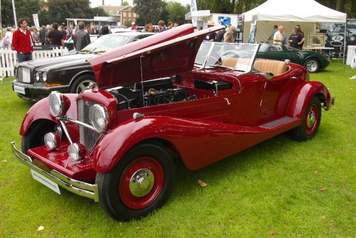 Pistonheads - The image captures a scene of a car show, where a striking red and silver vintage car is the center of attention. It's parked on a vibrant green field, surrounded by a few more classic cars and patrons admiring them. A man, presumably the car owner or show organizer, stands next to it, while under a white tent, several people are engaged in conversation. The vintage cars and the crowd create a lively atmosphere, highlighting the appreciation for classic rides in this setting.