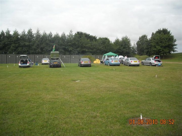 Pistonheads - This image depicts an outdoor scene on a day with overcast skies. There are several cars parked in a grassy area. They are of different colors and sizes, and some are covered with what appears to be tarpaulins or homemade covers. Each car has a smaller flag pole in front of it, and several flags are hanging from these poles. In the background, there are trees and a fence behind the grassy area where the cars are parked. The image has a watermark that reads "01.06.2010 08:52." The scene suggests a gathering or event where these cars may be on display.