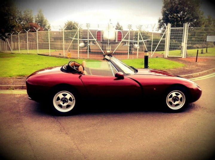 Show us your SIDE! - Page 22 - Readers' Cars - PistonHeads - This image depicts the front of a two-seater sports car with a vibrant red hue, parked on a street. The car appears to be small with two doors and is positioned in the foreground with a clear view of its front license plate area and part of its windshield. A wire fence is visible in the background, suggesting a roadside view. The car's design suggests that it is an open-top or convertible model, likely intended for a driver and passenger. The photograph is taken during the day under bright lighting conditions.