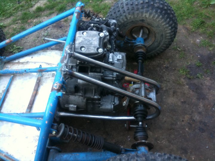 Kart Engined Diff Cross Pistonheads Bike - The image features a reclining racing kart, painted in a faded blue color, with a metallic frame. The kart's engine, mounted on the frame, appears to be in disarray, with several gears and components surrounding it. The wheels of the kart, which are particularly noticeable, are coated in a layer of mud, indicating recent outdoor use. Grass is also visible beneath and in those areas, further reinforcing the outdoor setting of the image.