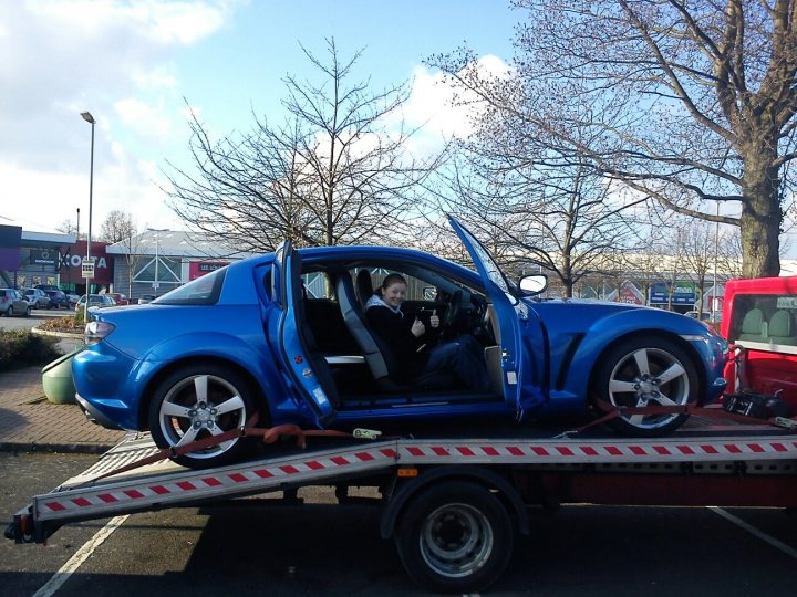 Mazda RX8 V8 1UZ-FE engined project, Insurance woes - Page 1 - General Gassing - PistonHeads - In the image, there's a vibrant blue sports car on a flatbed tow truck. A person is seated in the driver's seat, and the car is situated on the flatbed of the truck. The tow truck is parked in front of a store, with some other cars visible in the background. The sky above appears to be a clear, sunny day, adding a bright contrast to the scene.