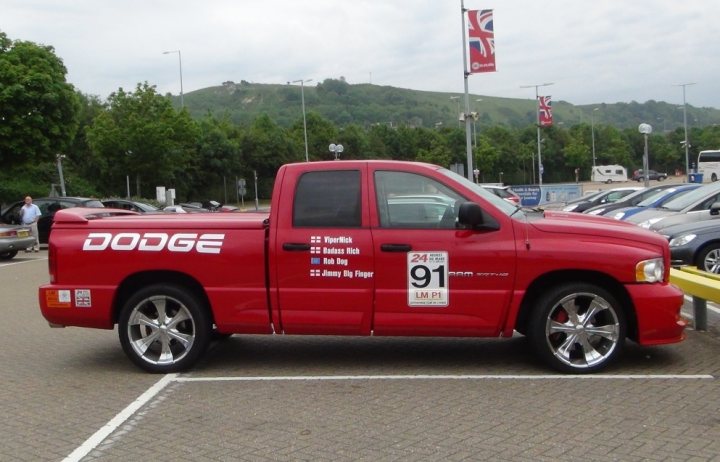 RAM at tunnel for Lemans trip - Page 1 - Vipers - PistonHeads - The image shows a bright red Dodge truck with some texts and numbers on the side, parked in a parking lot. In the background, there are other vehicles parked and a row of green trees. Additional elements in the image include a flag in the distance displaying the Union Jack, a bus on the right side, and a person on the left side of the image. The sky is overcast, suggesting it might be a cloudy day.