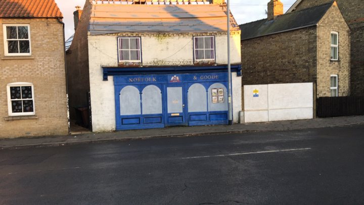 Funny/ridiculous company names - Page 1 - The Lounge - PistonHeads - The image displays a quaint, blue-painted, two-story building with a vibrant contrast to the rest of the scene. The upper portion of the building is peeling, hinting at its age, and there's visible damage on its side. In front of the building, a fenced yard extends onto the sidewalk, which is located alongside the structure. The scene gives an impression of a quiet, older neighborhood, possibly in a part of the United Kingdom where such buildings are common.