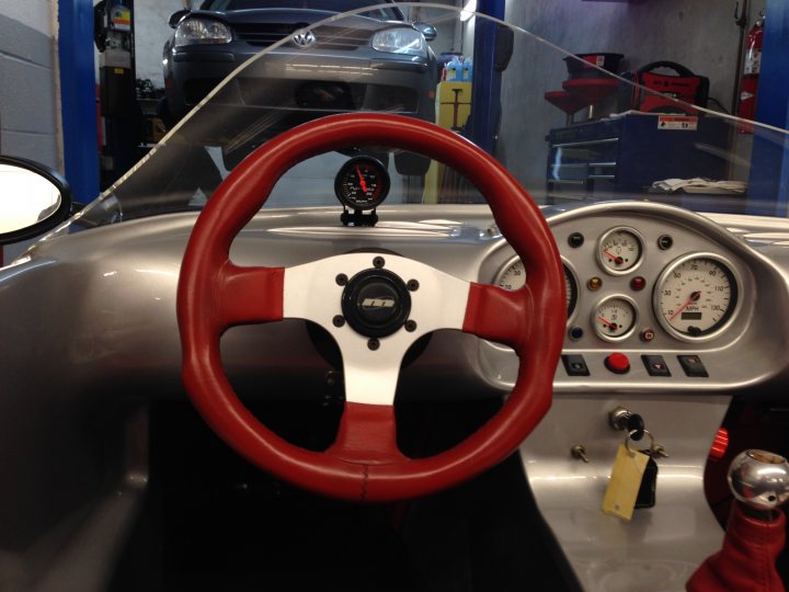 US G20 at track day - Page 1 - Ginetta - PistonHeads - The image captures the interior of a vehicle, with the focal point on the steering wheel. The steering wheel appears to be a red adhesive type with a white background and black switchgear. It is positioned at the center of the vehicle, equipped with a multitude of gauges and indicators that are typical of a race or performance car. To the side of the steering wheel, there is a prominent array of mechanical components that could be the engine or part of the vehicle's controls. The car itself is actually a picture of a car displayed in a shop window, suggesting that the viewer might be looking at a decal, sticker, or a realistic drawing meant to simulate the car's interior. The shop window is visible, hinting at a commercial setting.