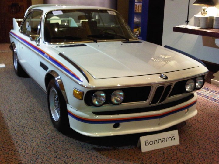 Bonhams Dubai Mirage Auction Pistonheads October Royal - The image showcases a classic vintage car on display, known as a BMW Bonhams R100GS, parked inside a building that could be a car show or a dealership. The car's body is white with silver and blue stripes along the side, resembling a racing design. Its headlights, tail lights, and wheels also appear sleek and in good condition. A small sign is placed in front of the vehicle, likely providing information about the car for visitors. Behind the car, the backrest of a chair is visible, suggesting that the car is positioned in a manner that allows people to move around it.