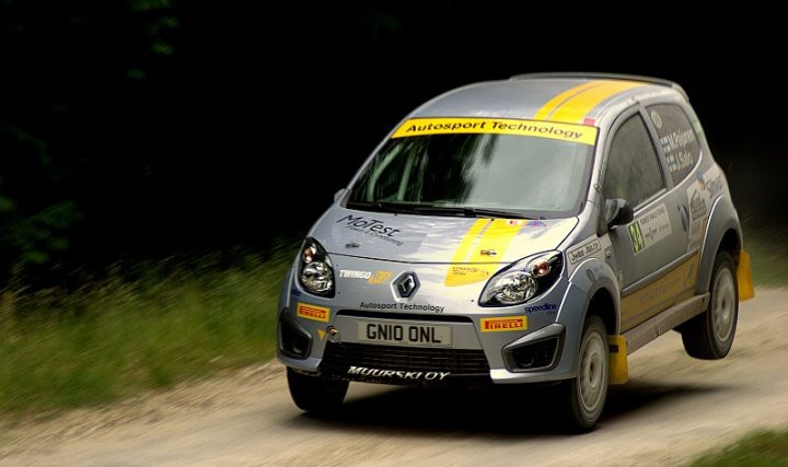 FOS good photography vantage points.. - Page 1 - Goodwood Events - PistonHeads - The image depicts a Renault Megane vehicle with a classic racing style, featuring distinct black and yellow stripes running along its body. The car is in motion on a dirt or gravel road, which is bordered by vegetation that suggests the photo was taken in a rural or semi-rural setting. The bright lighting indicates that the photo was likely taken during the day. The Renault Megane RS is explicitly visible on a placard on the side of the car, which suggests that this vehicle is a Renault Sports model, known for its high-performance capabilities.