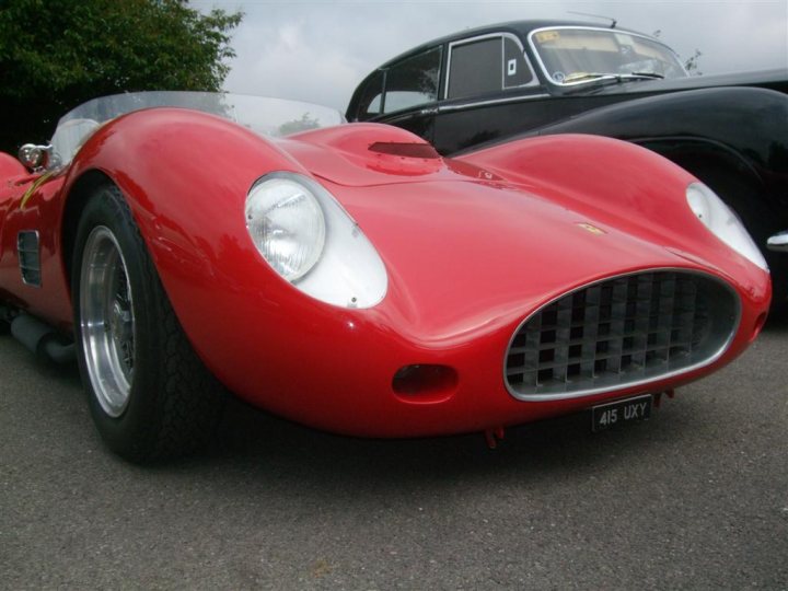 GRRC Open Day - 11 August - Page 1 - Goodwood Events - PistonHeads - The image shows a classic red sports car with a shiny finish, parked on a concrete surface with a black car in the background. The sports car features streamlined bodywork, large headlights flanking a prominent emblem, and a slanted design that contributes to its aerodynamic appeal. The black car behind it has a vintage design, possibly from the same era. Both cars are sleek and polished, reflecting light and suggesting a well-kept condition.