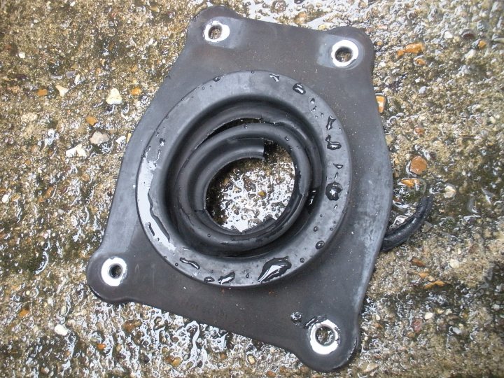 Miles Turret Change Years Pistonheads Oil - The image shows a black automotive rubber mount, typically used to isolate engine vibrations. The mount is lying flat on a wet, gravelly surface, and it appears to be wet with grease or oil, suggesting it may have been removed from a vehicle for servicing or replacement. The mount's unique curved shape, with a central circular protrusion, is designed to fit snugly over a steel or refractory ring, creating a flexible joint.