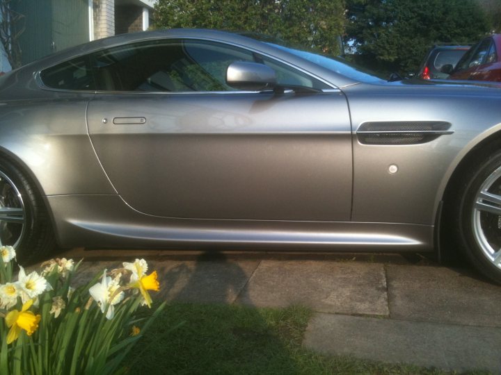 Slinc Pistonheads - The image shows a silver Aston Martin car parked on a driveway with neatly trimmed grass in the foreground. The car has a shiny, metallic finish and the brand's logo is visible on the hood. Behind the car, there's a house with a simple design. Off to the right, another car can be seen, suggesting that the Aston Martin is not alone in this driveway.