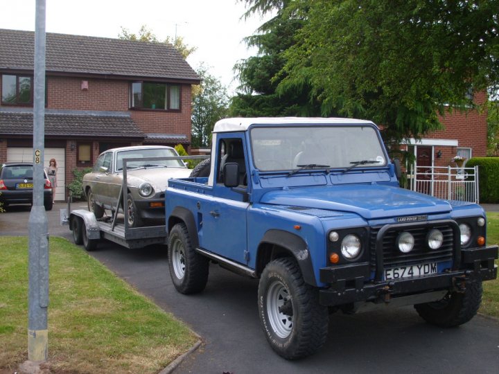 Pistonheads - The image shows a towing scenario where a vintage blue Land Rover Defender is either pulling or being pulled by a vintage silver car on a street lined with houses, trees, and a blue truck. The Land Rover has a flatbed and is carrying the small silver car in that manner. The tow is taking place, and the presence of multiple vehicles suggests a sense of scale and enterprise for the tow truck.