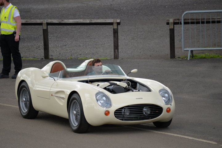 Spotted 2017 - Page 13 - North East - PistonHeads - The image features a man riding in a vintage sports car on a street. The car is white and appears to have a convertible top down, revealing the interior. The vehicle is moving along a road and is facing towards the left side of the image. The driver is seated in the car, attentively observing the road. There's also a blurry figure on the left side of the photo, suggesting the presence of someone in yellow, possibly a traffic controller or roadside assistance. The overall setting seems to be a city street or a rural road.