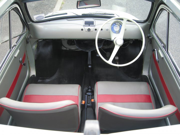 Fiat 500 - Page 1 - Readers' Cars - PistonHeads - The image shows an interior view of the passenger side of a convertible car. The steering wheel is positioned on the left side of the vehicle, which indicates that it's designed for a driver in the Right (British-style) lane, as opposed to the Left (American-style) lane. The interior of the car has a two-tone gray color scheme with red accent piping and matching red cushion covers. The vehicle appears to be parked with the top in the down position. Since the photograph is taken from the passenger side, the gear shift lever's position is visible near the top of the image.