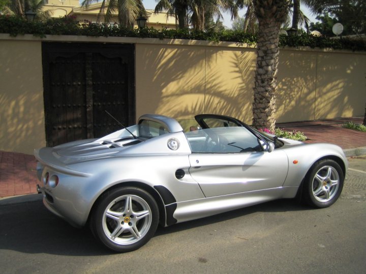 A black and white photo of a car parked in a parking lot - Pistonheads - The image showcases a sleek silver sports car parked on a street. The car is positioned in front of a beige stucco wall with a decorative wooden gate. A palm tree is visible to the right of the frame, creating a tropical ambiance. The car, with its glossy exterior and a soft top roof, seems ready for a leisurely drive.