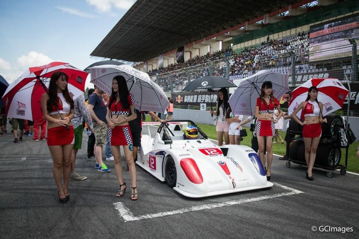 My new Radical in China... - Page 1 - Radical - PistonHeads - The image captures a scene at a race track event, with several young women trotting around a white race car, each carrying a matching open umbrella. They are dressed in coordinated red and white outfits, mirroring the team's colors. In the background, a crowd of spectators can be seen, indicating that this is a public racing event. The focus of the image is the interaction between the girls, the car, and the umbrellas, creating a visually coordinated and dynamic scene.