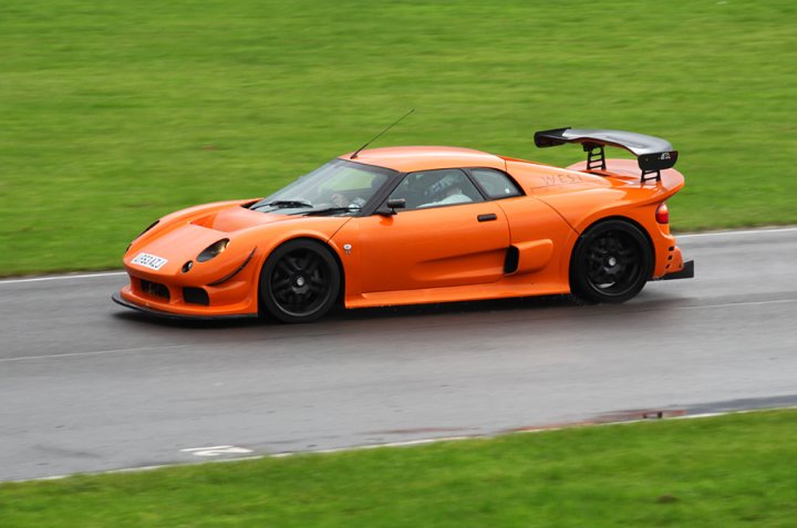 Pistonheads - The image features an orange sports car with a large rear wing, driving at speed down a paved roadway. The car's sleek design and vibrant color suggest it's built for high-performance driving. The setting appears to be a rainy day, as hinted by the wet roads and the car's aerodynamic profile with hints of water droplets. The experience of the car implies that it's tailored for a thrilling ride.