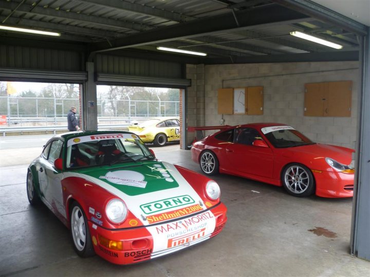 14th Annual Oulton Park RS day - Page 6 - Porsche General - PistonHeads - The image presents a spacious garage housing three distinct cars. In the forefront, a red and green Porsche, bearing the number 3, takes center stage. Parallel to it, a yellow and white Porsche is parked, while a red and white Porsche, also with the number 3, is situated behind the first car, close to a sign advertising "Torno Shelahworld 9PM". On the left, a person is standing next to the Porsche with "Klippo" written on it, possibly observing or maintaining the vehicles. The garage's structure allows for ample space between the parked cars, suggesting it is designed for multiple vehicles.