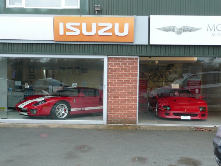 Pistonheads Ford Monks Heath Sale - The image shows the front of an automotive dealership named "Isuzu". Inside the glass display area, two red sports cars and a motorcycle are prominently displayed. The vehicles feature streamlined designs with sporty accents, indicative of high-performance models. The dealership's exterior facade includes brick elements and the manufacturer's logo at the top. The setting appears to be a typical urban environment.