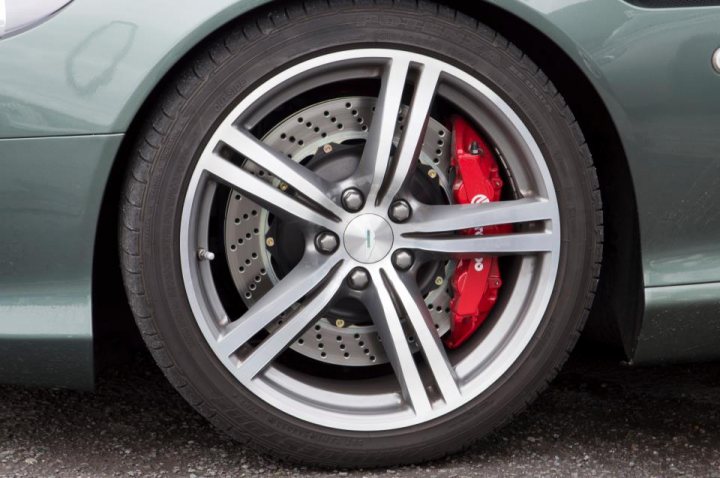 Brakes - Page 1 - Aston Martin - PistonHeads - The image shows the front wheel of a green car, likely taken from a certain angle or perspective. The wheel is black with shiny silver rims giving a contrasting look. The center hubcap is attached to the hub, which has a distinctive red label on it. The background of the image is silver and metallic, keeping the focus on the wheel and car. The overall condition of the car and wheel appears to be smooth and well-maintained.