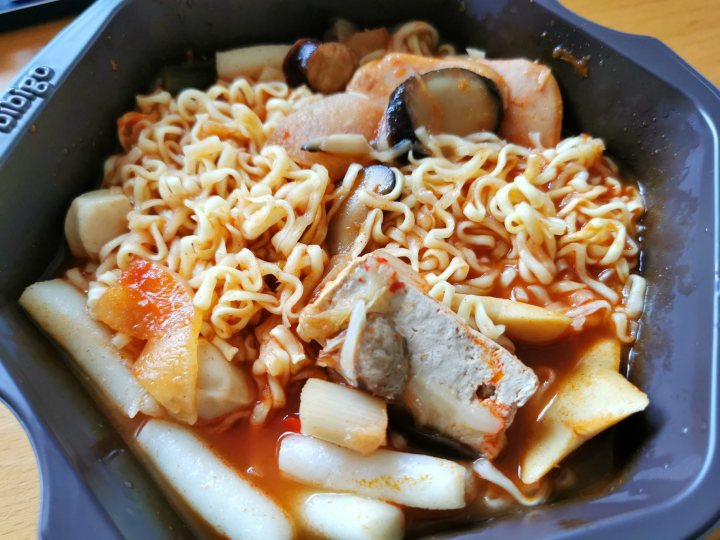 A plate of food with meat and vegetables - Pistonheads - This image features a tray of food on a table. The main components of the meal are noodles and meat, possibly beef. The noodles are topped with a sauce that looks like it could be tomato-based, and there's an additional sauce or condiment on top. To the side, there's a small portion of what appears to be pickled vegetables. A can of instant noodles is visible in the tray. The meal is served in a styrofoam container, suggesting it might be takeout food. There's a spoon resting in the middle of the tray, ready for use.