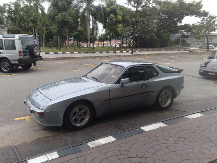 Pistonheads - This image features a gray sports car parked on the street. The car is sleek and compact, with a prominent rear wing and side air vents. It's positioned across two parking spaces, as indicated by the dashed lines on the concrete. In the background, there is a lush green environment, including trees, a grassy area, and another vehicle further away. The overall scene suggests an urban setting with public parking available.