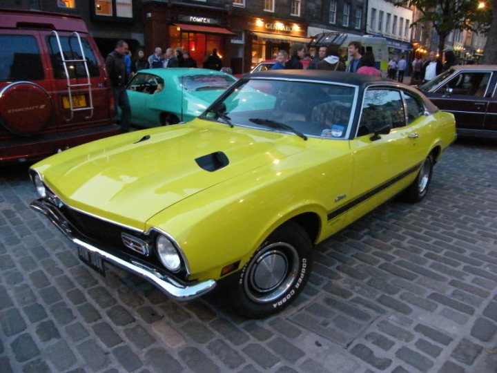 Cruisemeet Pistonheads Edinburgh - The image features a vibrant scene of a vintage car parade on a busy city street. The car in the spotlight is a classic yellow Mustang convertible, glossy and full of character, parked amidst other classic cars. The cars are parked adjacent to each other, with the Mustang prominently displayed. In the background, there are a few people and stores, indicating a lively urban atmosphere. A red van is partially visible in the background, adding to the dynamic street scene.