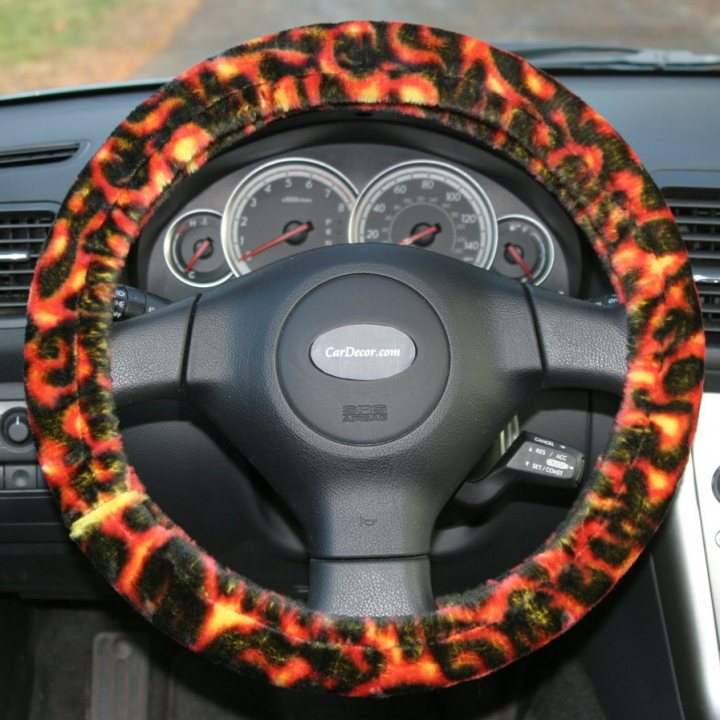 Steering wheel upgrade?? - Page 1 - Elise/Exige/Europa/340R - PistonHeads - The image depicts a car steering wheel with a cheetah print skin cover. The skin has prominent cheetah spots in a range of vibrant colors including orange, yellow, and black. The steering wheel is attached to a car dashboard, which is not fully visible in the photo. The steering wheel is black in color, contrasting with the colorful skin, and has the brand Cadillac logo imprinted largely in the middle. In the background, part of a sideview mirror with a material interior is visible.