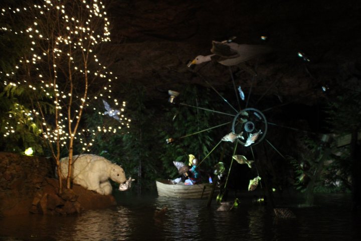 Santa in a cave  - Page 1 - South West - PistonHeads - The image captures a magical night scene. A Christmas tree stands sentinel on the left, its branches adorned with numerous white lights. In the heart of the scene, a wooden boat is gently floating on still water, its tranquility disturbed only by the swirling snowflakes that dance in the air, their light falling like stars. To the right, a whirlwind of celebratory lights decorates a large tree, conjuring a festive atmosphere. The entire scene is bathed in the ethereal glow of colored lights, creating a stark contrast with the peaceful water and the solitary white bear statue that sits on the banks, quietly observing the spectacle.