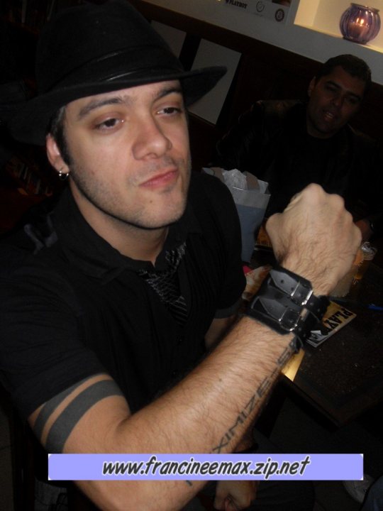 The image shows a man sitting at a table in a room with a low ceiling. He is wearing a black shirt, a black hat, and a skinny black tie. On his wrist, there is a black strap with a gold clasp fastened. His gaze is directed off to the side, and he has a serious expression on his face. In the background, another person is partially visible but not the focus of the image. Additionally, the website "www.fancineemax.zip.net" is overlaid on the image, suggesting that this is a filtered or edited photo.