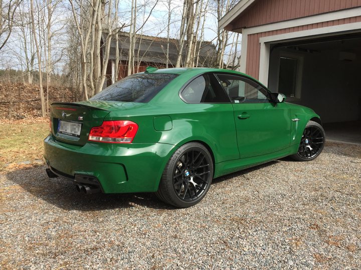 BMW 1M in dark green  - Page 1 - Readers' Cars - PistonHeads - The image features a striking green or possibly blue car parked in a driveway next to a house. The car is positioned at an angle, with the front of the car facing towards the right side of the frame. It has a sleek and modern design, with black rims accentuating its appearance. The driveway is surrounded by trees and rocks, giving the scene a natural and serene atmosphere. The car's vibrant color contrasted against the more muted tones of the driveway and surroundings create a visually striking image.