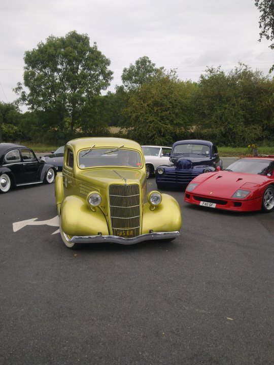 Pistonheads - In the image, a collection of classic cars is parked on a blacktop surface. Among them is a striking yellow car with a chrome grill, and a distinctive red bug with a pointed nose. Appliances and modern-day vehicles are in the background, suggesting a blend of antiquity and modernity. The scene appears to be outdoors during the day under cloudy skies. The cars are arranged facing the same direction, giving a sense of order amidst the variety of vintage models.