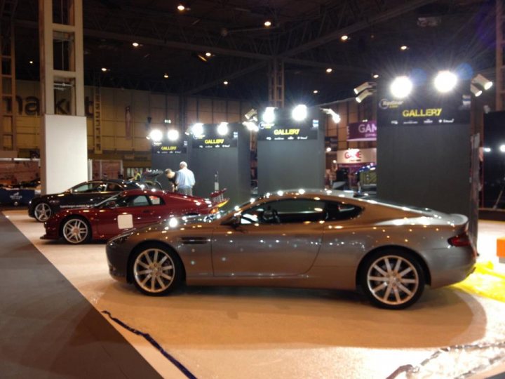 Top Gear Live (NEC this weekend) - Page 1 - Aston Martin - PistonHeads - The image captures a scene from a car show where several luxury cars, featuring a mix of red and gray colors, are elegantly displayed on a carpeted floor. The setting implies a circular or semi-circular layout, with the cars placed on pedestals, showcasing their design and quality to the viewers. The illumination suggests that the venue is well-lit, enhancing the visual appeal of the vehicles. The audience is present, with several people engrossed in observing and appreciating the cars on display.