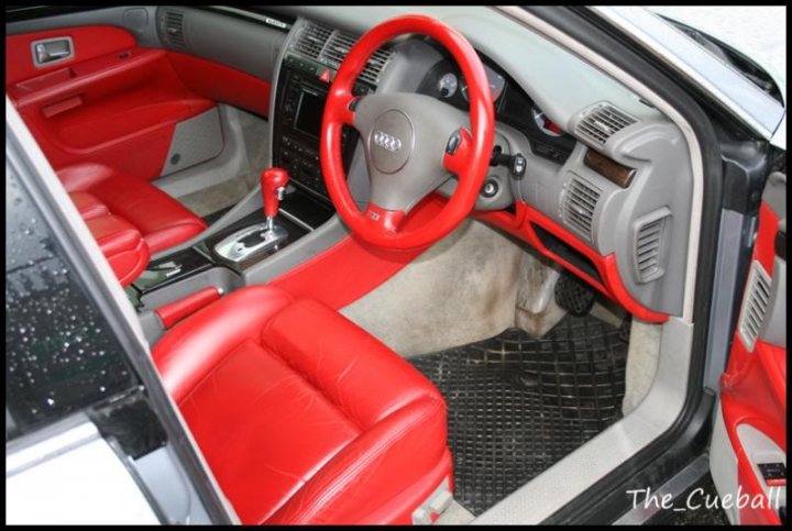 Can a dodgy interior put you off a car? - Page 2 - General Gassing - PistonHeads - The image is a photograph of the interior of a vehicle, focusing on the driver's side. The car appears to be a convertible due to the visible part of the roof that is retracted. The visible section of the vehicle is dominated by a striking combination of red leather upholstery and a grey plastic interior panel. The driver's seat is in the folded-down position, revealing that the vehicle is equipped with a manual transmission. There is no person visible in the photo. The steering wheel is also separated from its usual position, drawn down and resting on the dashboard. The image has a logo and text in the bottom right corner that reads "The Cubeball". The style of the image is straightforward, capturing the unusual view of the interior without any added embellishments.