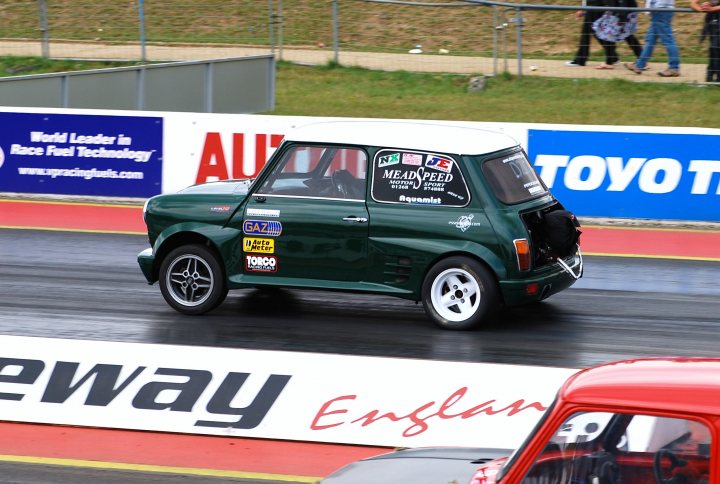 Pistonheads Park Mini - This image depicts a small green race car on a racetrack. The car is adorned with various stickers on the windows and door, showcasing names and logos, indicative of sponsorships. The tires are black and appear to be made of soft compound material, suitable for racing grip. The car is halfway around a corner, with a white curb on its right. The background features red and yellow caution fencing and other colorful banners advertising different companies. Visible spectators are enjoying the action from behind a blue barrier fence. The overall scene conveys the excitement and competition of a motorsport event.