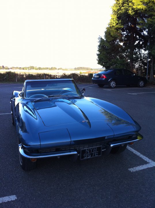 Thames Night July Valley Pistonheads Wednesday - The image shows a vintage sports car parked at an angle in a parking lot. The car is predominantly blue with a visible logo on the driver's side portion. It stands out against the backdrop of a bright sky. Adjacent to the car, there are parked vehicles, including another car and a dark SUV. The setting appears to be a rural area, as indicated by the open space in the background and the presence of a line of tall trees.