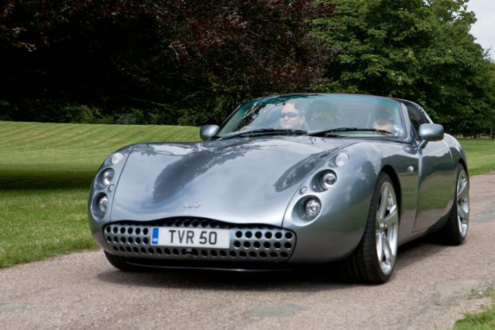 Motors Manor Pistonheads - The image captures a man driving a silver vintage car, which has a distinctive design featuring a prominent grille with vertical bars. The car is a two-seater convertible, as suggested by the convertible top and the absence of additional passenger doors. The vehicle bears a TVR 50 license plate, indicating that it might belong to a collection or special interest group of cars. The car is parked alongside a road lined with well-maintained grass and trees, suggesting a peaceful suburban setting.