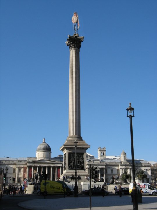 The London Pub crawl....................... - Page 1 - Vipers - PistonHeads - The image features a monumental pillar with a statue perched at the top. The pillar stands prominently against a clear blue sky, with the statue positioned on a cube-like platform. Below the pillar, several people are walking around, and there are cars visible in the background. The overall scene suggests a public square or city park during the daytime.