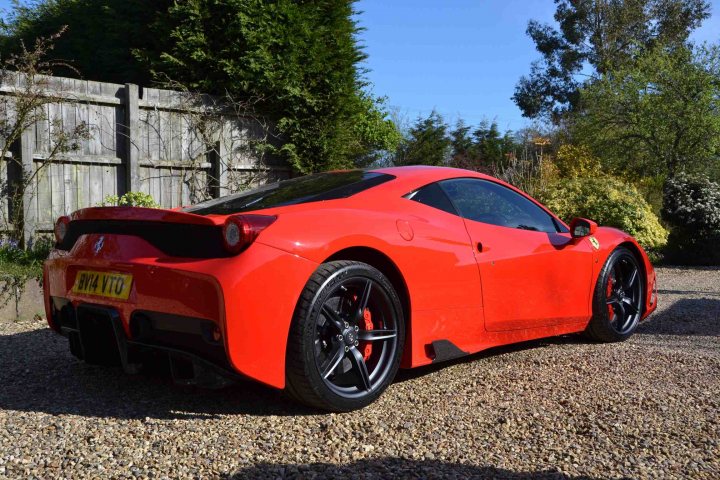 458 Speciale - Page 1 - East Anglia - PistonHeads - The image presents a vibrant scene featuring a mid-engine sports car, painted in a striking shade of red. The vehicle is parked on a gravel lot, adjacent to a weathered wooden fence and lush trees. The driver's side door is open, suggesting the car is being accessed or prepared for a journey. The setting seems peaceful and serene, with the car's glossy finish contrasting beautifully against the natural surrounds.