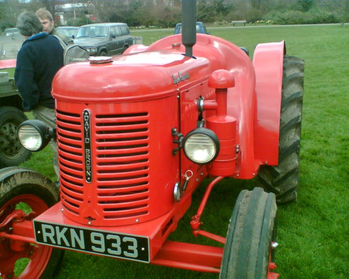 Our Heritage - Page 1 - Aston Martin - PistonHeads - In the image, a large red tractor with the registration number RKN 933 is the central focus. It's parked on a grassy area, its front face directly into the camera. There is a visible PTO shield on the front of the tractor. The background reveals multiple other vehicles, including cars and a truck, indicating that this might be a parking area for various vehicles. A bench is also visible in the distance, adding to the outdoor setting of the scene.