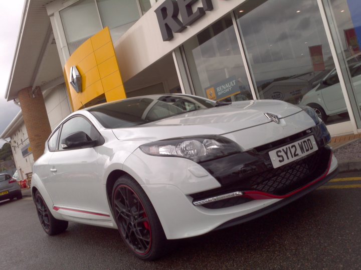 A car parked next to a parking meter - Pistonheads - This image shows a white coupe parked in front of a car dealership. The dealership has a large glass window featuring the Renault logo. To the right, there is a white Fiat van parked in front of the store. The ground appears to be wet, suggesting recent rain. The vehicle in focus has red accented rims, adding a pop of color to its design.