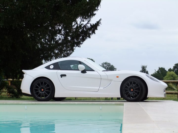 Pistonheads - The image showcases a sleek, white sports car positioned at an angle near the edge of a rectangular swimming pool on a sunny day. The car, which appears to be quite modern, has a polished and well-maintained appearance. Leaning slightly over the pool, the photographer captures a reflection of the vehicle on the pool's surface. In the background, a calm marine environment is visible with distant trees and bushes under a hazy sky.